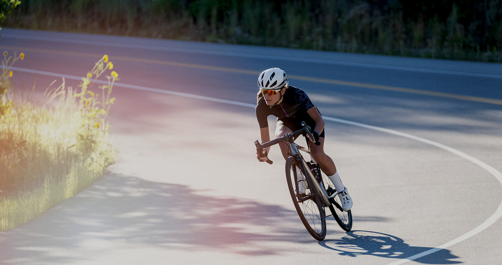 A female cyclist pedals around a corner going downhill wearing black clothing and a white helmet.
