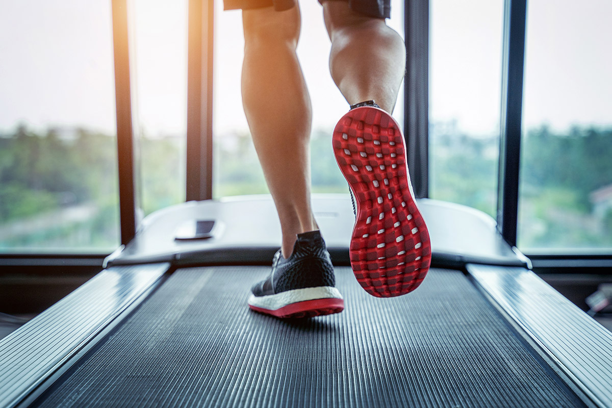A close up of a man's legs running on a treadmill wearing black and red running shoes looking out a gym window.